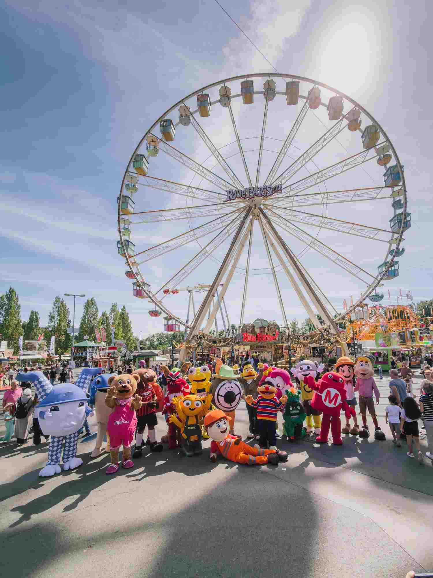 20260708-IMG_5449-klein Maskottchen der Maskottchenparade auf dem Schützenplatz vor dem Riesenrad. Zu sehen sind: Rasti, wali, Lu, Ernie und Bert, Hektor, orangener Müllmann, HTpino, Makey und Botti, Infalino, Follow-Bee Frida, Hajo, Narri, Idex, Eddi, Hosenwackler und Dame