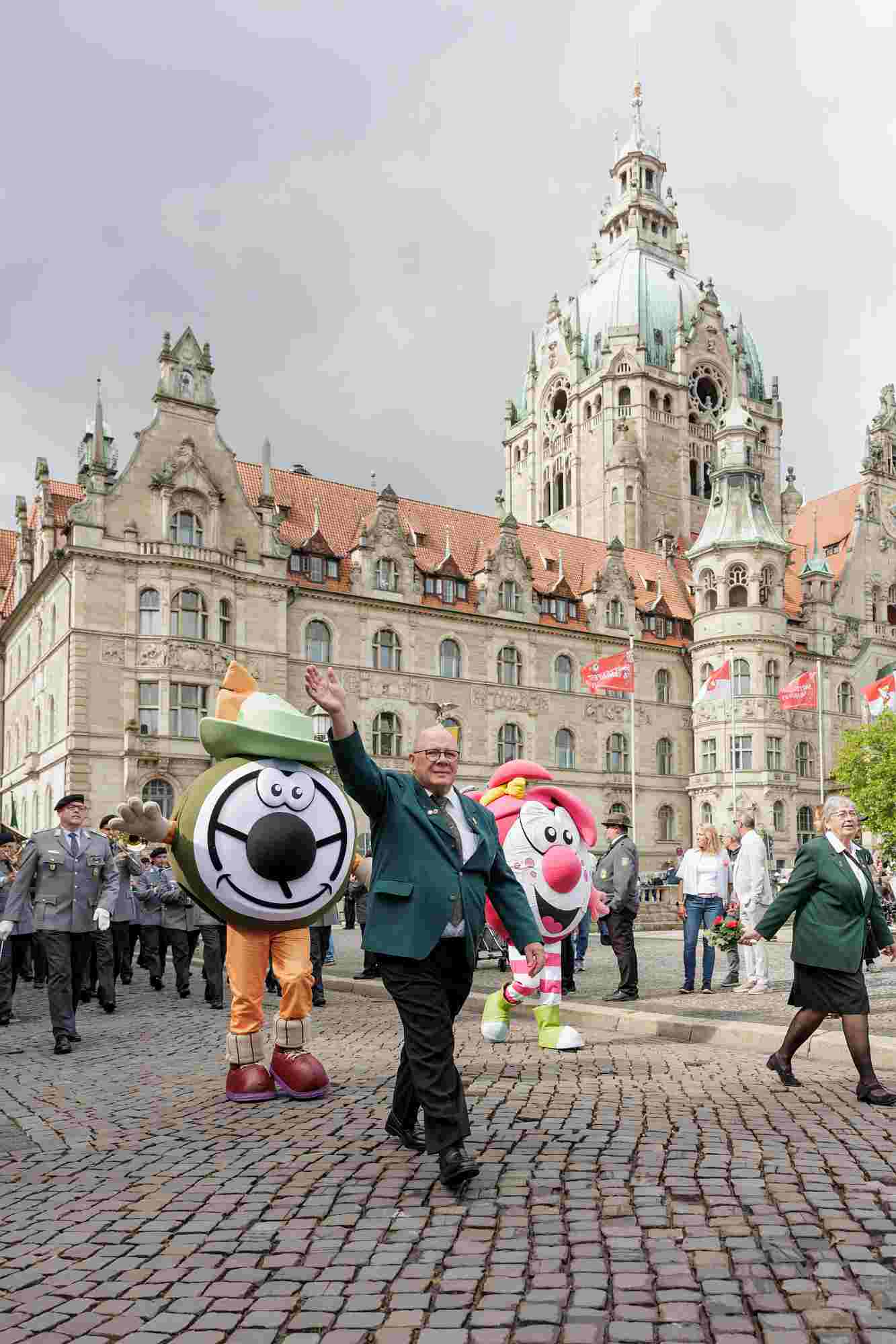 Schützenfest 2026 Im Vordergrund laufen die beiden offiziellen Werbefiguren des hannoverschen Schützenfestes mit, links Ballerkalle und rechts daneben Ballerina. Paul-Eric Stolle in grüner Schützenjacke winkt fröhlich. Im Hintergrund zu sehen ist das neue Rathaus.