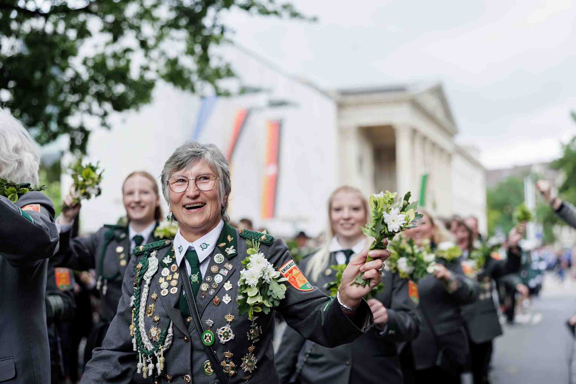 Schützenfest 2026 Im Mittelpunkt steht eine freudig lächelnde Schützin des Schützenvereins Engelbostel in traditioneller Festuniform. Sie trägt zahlreiche Ehrenbegegnungen, Abzeichen und Orden auf ihrer Jacke und hält, genau wie die anderen Marschierenden im Hintergrund, einen kleinen Blumenstrauß in der Hand. Der Festumzug bewegt sich durch die Straßen Hannovers, vorbei an großen Flaggen im Hintergrund