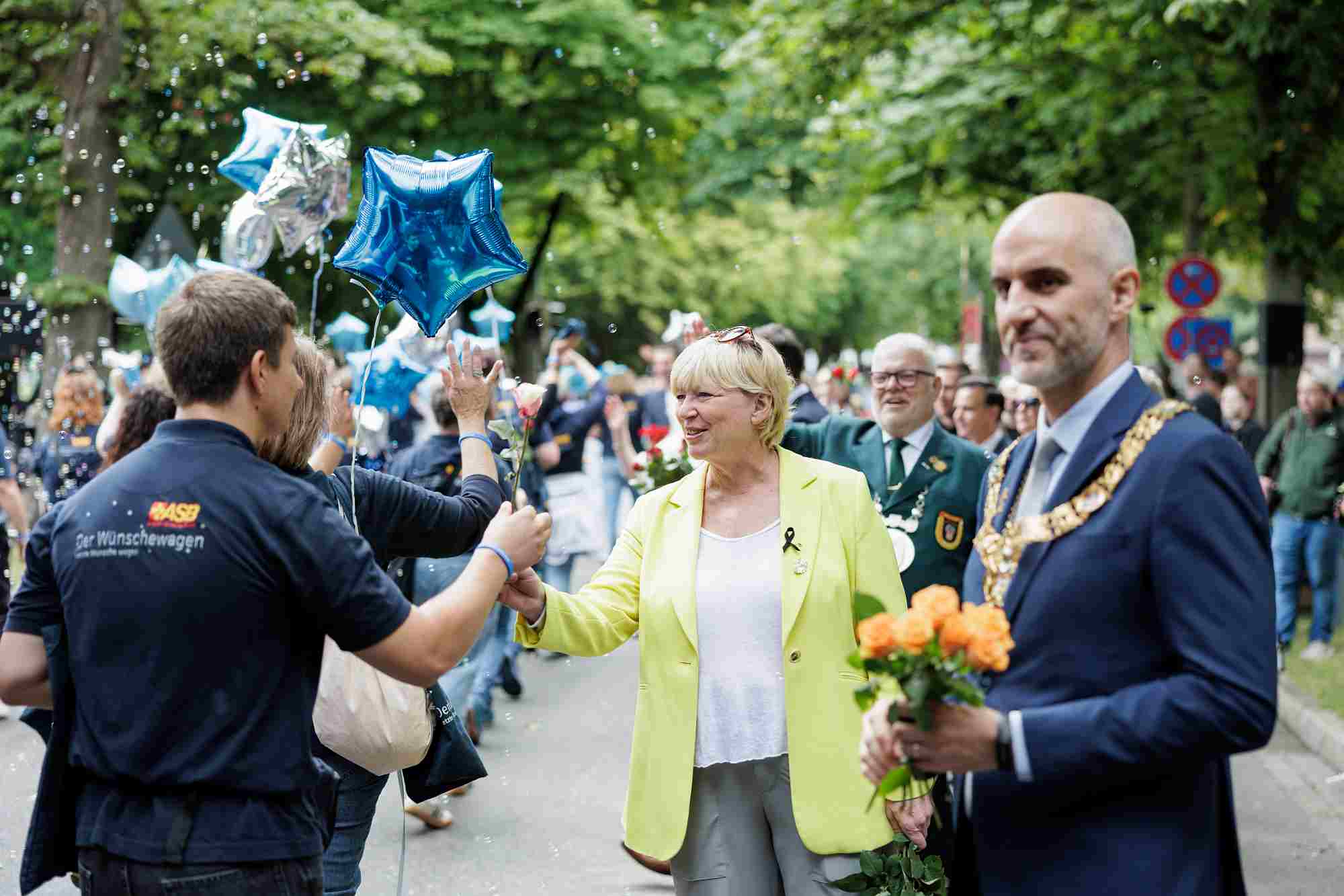 Schützenfest 2026 Auf dem Bild sind Hannovers Oberbürgermeister Belit Onay und die niedersächsische Regionalministerin Wiebke Osigus beim traditionellen Schützenausmarsch in Hannover zu sehen. Belit Onay hält einen Strauß orangefarbener Rosen in den Händen. Wiebke Osigus nimmt lächelnd eine einzelne Rose von einem Mitarbeiter des ASB (Arbeiter-Samariter-Bund) entgegen, der ein T-Shirt mit der Aufschrift „Der Wünschewagen“ trägt.
