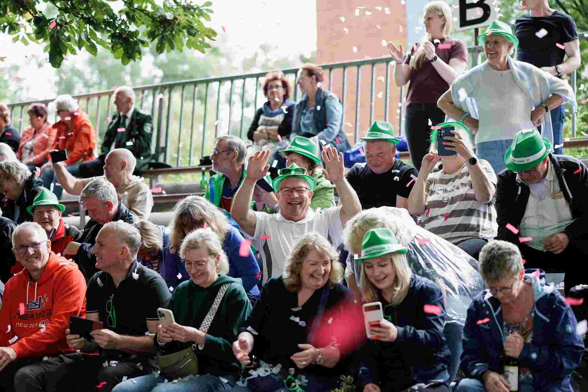 Schützenfest 2026 Verschiedene Personen sitzen auf einer Treppe oder Tribüne und tragen grüne Hüte, einige halten Smartphones hoch oder fotografieren das Geschehen, während Konfetti durch die Luft fliegt.