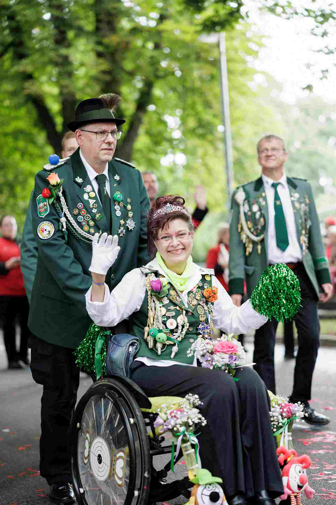 Schützenfest 2026 Zu sehen ist eine Schützin im Rollstuhl, sie trägt eine weiße Langarmbluse, kombiniert mit einem hellgrünen Halstuch, über der Bluse trägt sie eine dunkelgrüne Weste, die prunkvoll mit zahlreichen Schützenorden, Ehrenzeichen, Anstecknadeln und Plaketten besetzt ist und eine funkelnde Tiara im Haar. Sie trägt weiße Festzeithandschuhe und hält in der linken Hand einen grünen Pompon. Ein kleiner Blumenstrauß aus rosa und weißen Rosen liegt auf ihrem Schoß. Ihr Rollstuhl ist festlich mit Blumenarrangements an den Seiten geschmückt. er Schütze hinter dem Rollstuhl:• Uniform: Er trägt den klassischen grünen Schützenrock (Uniformjacke) mit dunklem Kragen. Am Revers befinden sich Vereinsabzeichen und an der linken Brusttasche mehrere große Orden und Ehrenzeichen. Der Schütze hinter ihr, trägt auf den Schultern Epauletten. Zudem verläuft eine silberne Schützenschnur quer über die Brust. Auf dem Kopf trägt er den traditionellen grünen Schützenhut, der mit einer typischen Feder verziert ist.