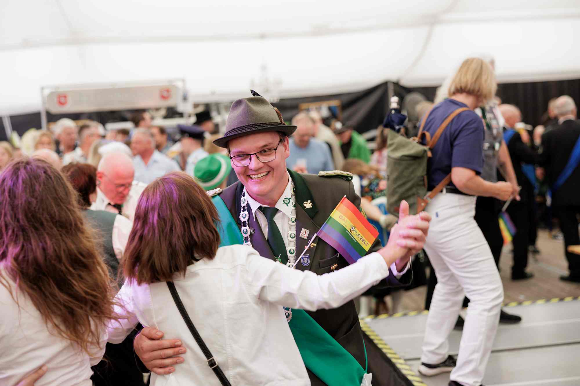 Schützenfest 2026 Das Bild zeigt fröhliche Menschen in traditioneller Schützenkleidung und Tracht, die in einem Festzelt tanzen und eine kleine Regenbogenfahne schwenken.