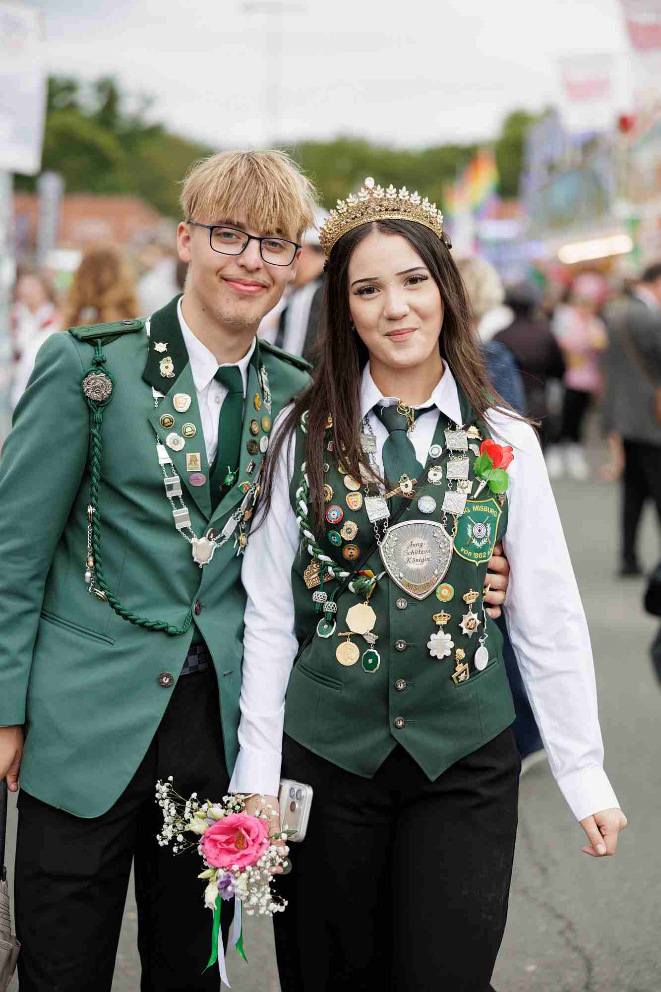 Schützenfest 2026 Auf dem Bild sind zwei junge Mitglieder eines traditionellen deutschen Schützenvereins beim Schützenfest Hannover zu sehen. Auf dem runden Ärmelabzeichen der Frau steht „USG Misburg von 1862“ und auf ihrem großen Brustschild ist der Titel „Jung-Schützen Königin“ zu lesen. Es handelt sich hierbei um die amtierende Nachwuchs-Majestät des Vereins und ihren Begleiter.