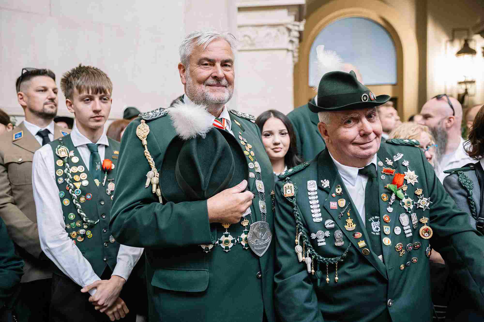 Schützenfest Hannover 2026 Im Bild stehen Männer in traditioneller Schützenuniform, dazu gehören Klassische, dunkelgrüne Schützenjacken und aufwändige Epauletten mit Kordeln und Fransen, die den jeweiligen militärisch inspirierten Rang innerhalb des Vereins anzeigen. Die Vorderseiten der Uniformen sind dicht mit zahlreichen Schützenorden, Ehrenzeichen und Anstecknadeln bestückt, die von sportlichen Erfolgen oder langjährigen Mitgliedschaften zeugen. Der rechte Mann trägt den typischen Schützenhut mit einer Kordel und Vereinsabzeichen. Der linke Mann hält seinen Hut mit einem weißen Flaumbusch in der Hand