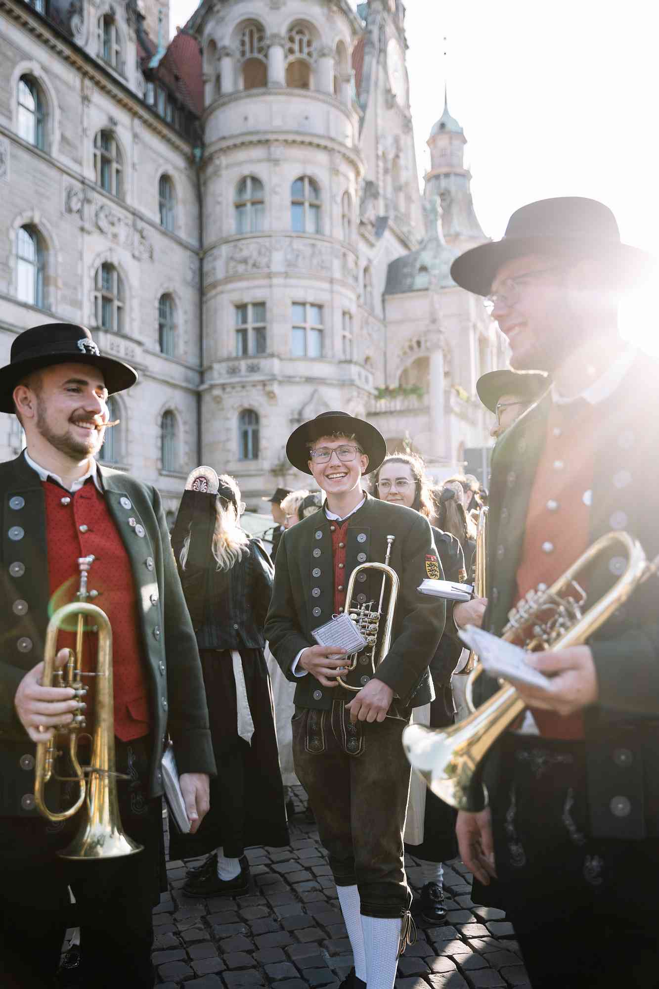 Schützenfest Hannover 2026 Zu sehen sind drei Musiker einer bayerischen Trachtenkapelle in typischer Tracht mit Lederhosen, Haferlschuhen, roten Westen und den Trachtenbehütungen vor dem neuen Rathaus in Hannover.